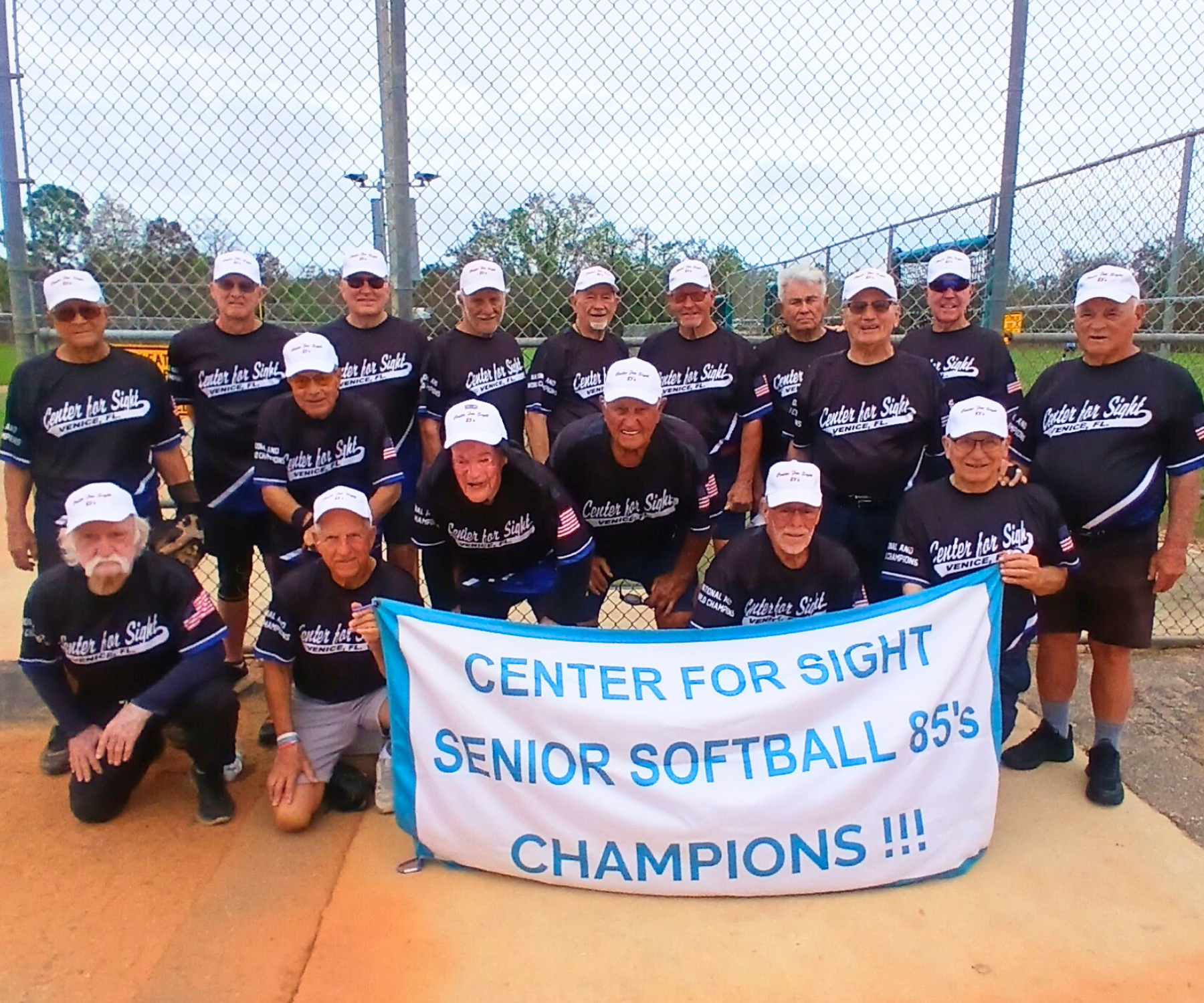 Group of senior softball players in matching uniforms posing with a banner reading 'Center for Sight Senior Softball Champions'.